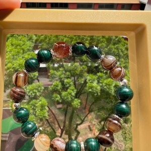 Op de afbeelding: Een groene, bruine en oranje kralen armband met zilveren accenten. De armband is gemaakt van natuurlijke stenen en wordt getoond in een doorzichtige doos.
