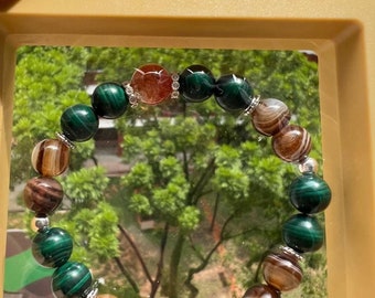 Könnte beinhalten: Ein grünes, braunes und orangefarbenes Perlenarmband mit silbernen Akzenten. Das Armband besteht aus natürlichen Steinen und wird in einer durchsichtigen Schachtel präsentiert.
