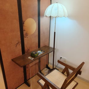 Op de afbeelding: Een houten console met een heldere acrylbasis, met boeken en een kleine doos. Een vloerlamp in vintage-stijl met een geschulpte kap staat in de buurt. Een houten stoel met een geweven zitting staat ook in de kamer. De achtergrond is een decoratief scherm.