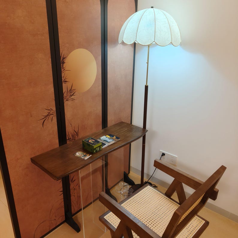 Op de afbeelding: Een houten console met een heldere acrylbasis, met boeken en een kleine doos. Een vloerlamp in vintage-stijl met een geschulpte kap staat in de buurt. Een houten stoel met een geweven zitting staat ook in de kamer. De achtergrond is een decoratief scherm.