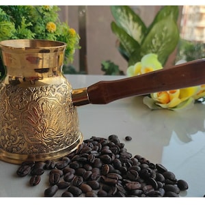 May include: A gold-coloured brass coffee pot with an ornate design and a wooden handle. The pot is sitting on a surface with coffee beans scattered around it.