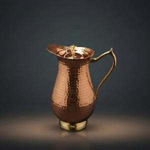 May include: A copper pitcher with a hammered texture, featuring a curved handle and a lid. The pitcher has a gold-coloured base and handle, and it is set against a dark background.