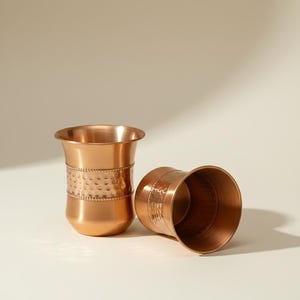 May include: Two copper-coloured metal cups. One cup is upright, and the other is lying on its side. Both cups have a flared rim and a textured band around the middle. The cups are on a light-coloured surface.