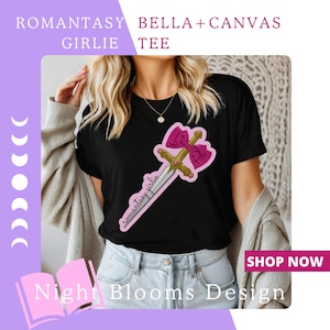 May include: Black t-shirt featuring a sword and pink bow graphic. The text "Romantasy Girlie Tee" is printed at the top. The words "Night Blooms Design" are at the bottom. The shirt is Bella + Canvas.