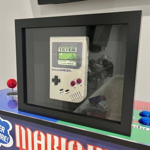 May include: A vintage white Game Boy handheld video game console with a black frame and a clear glass cover. The console is displaying the game Tetris on the screen. The console is framed and displayed on a wall.