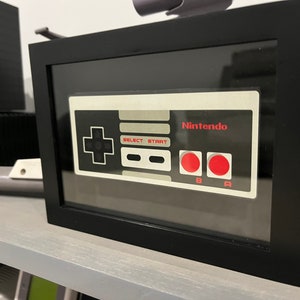 May include: A framed black and white image of a classic Nintendo NES controller with red buttons. The controller is in a black frame with a glass front. The text "Nintendo" is printed on the controller.