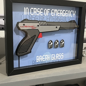 May include: A framed picture with a black frame and a blue background. The picture features a gray and red toy gun with the text "In Case of Emergency Break Glass" and "Nintendo" printed on it. Three gray bullets are also pictured.