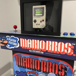 May include: A vintage Nintendo Game Boy handheld console with a black and white screen and a gray plastic case. The console is displaying the game Tetris. The Game Boy is framed in a black frame and mounted on a wooden stand. The stand is decorated with a blue and red Mario Bros. logo and a 2-player feature label.