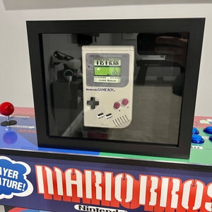 May include: A vintage white Game Boy handheld gaming console with a black and white screen displaying the game "Tetris". The Game Boy is framed in a black frame and is sitting on top of a blue and red arcade cabinet with the words "Mario Bros" printed on the front.
