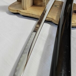 Pu&ograve; includere: Una katana giapponese tradizionale con una guaina laccata nera e una lama in argento lucido. La spada &egrave; esposta su un supporto in legno.