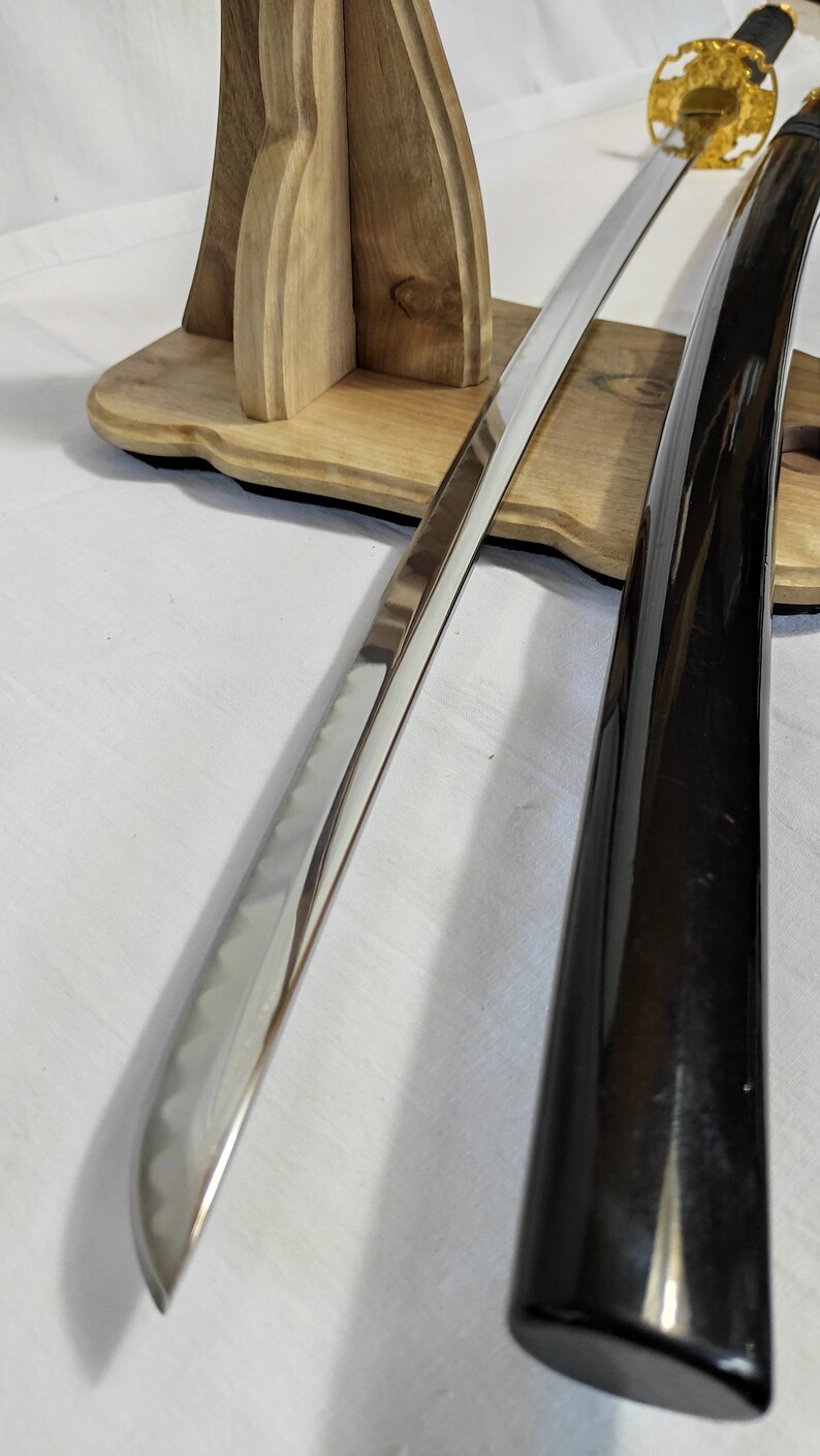 Pu&ograve; includere: Una katana giapponese tradizionale con una guaina laccata nera e una lama in argento lucido. La spada &egrave; esposta su un supporto in legno.