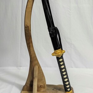 Pu&ograve; includere: Un supporto per spada in legno con un design curvo, che tiene una katana nera con un'impugnatura dorata e un'impugnatura avvolta in bianco e nero.