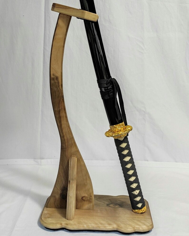 Pu&ograve; includere: Un supporto per spada in legno con un design curvo, che tiene una katana nera con un'impugnatura dorata e un'impugnatura avvolta in bianco e nero.