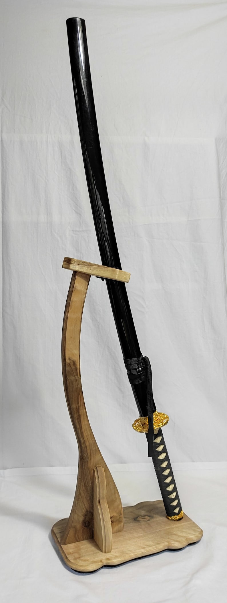 Pu&ograve; includere: Una katana nera con una tsuba (guardia) dorata e una saya (fodero) bianca e nera &egrave; esposta su un supporto in legno.