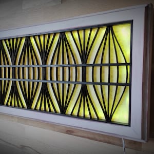 May include: A rectangular wall-mounted light fixture with a white frame and a black geometric design. The light shines through the design, creating a yellow glow.