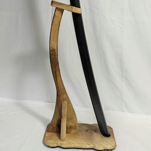 Pu&ograve; includere: Un supporto per spada in legno con un design curvo. Il supporto &egrave; in legno marrone chiaro e ha una spada nera appoggiata su di esso.
