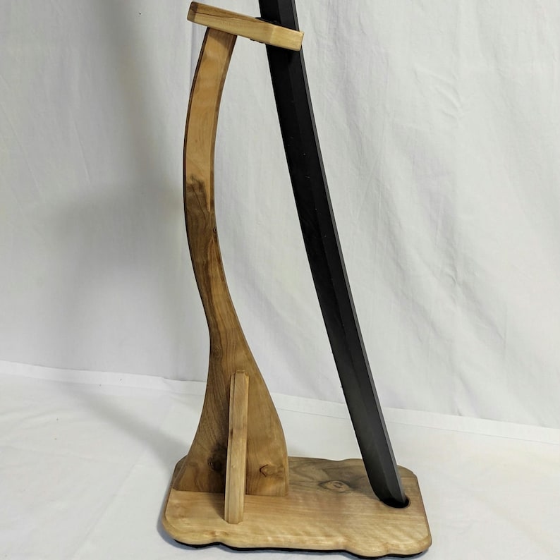 Pu&ograve; includere: Un supporto per spada in legno con un design curvo. Il supporto &egrave; in legno marrone chiaro e ha una spada nera appoggiata su di esso.