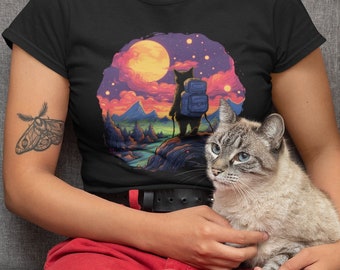 Puede incluir: Una mujer lleva una camiseta negra con un gráfico de un gato con una mochila haciendo senderismo en un paisaje colorido. El gato está de pie en un acantilado con vistas a un río y montañas. La luna y las estrellas son visibles en el cielo. La mujer está sosteniendo un gato en su regazo.