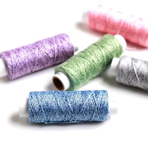Pode incluir: Uma coleção de bobinas de linha coloridas em várias tonalidades, incluindo azul, verde, rosa, roxo e cinza. A linha é enrolada firmemente em torno de tubos de cartão branco, criando uma aparência texturizada. As bobinas estão dispostas sobre um fundo branco.