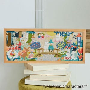 May include: A framed cross-stitch featuring a colorful scene with cartoon characters, a bridge, and floral elements. The artwork is set in a light wooden frame and rests on a stack of books. The image includes the text "©Moomin Characters™".