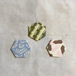May include: Three hexagonal ornaments with different patterns. One is light blue with a white floral design, another has a green and yellow camouflage pattern, and the third has a brown and pink cow print.