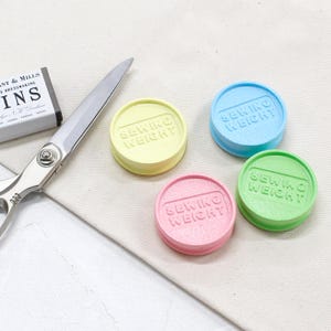 Puede incluir: Cuatro pesas de costura de colores pastel, amarillo, azul, rosa y verde, cada una con la inscripción "SEWING WEIGHT" en relieve. Unas tijeras plateadas y una caja de alfileres sugieren un tema de costura o artesanía.