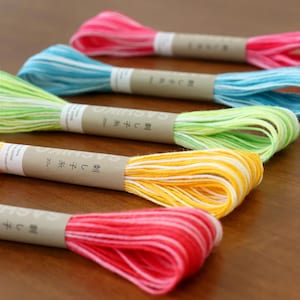 Puede incluir: Cinco madejas de hilo de bordar Sashiko dispuestas en fila. Cada madeja presenta una paleta de colores variados que incluye rosa, azul claro, verde lima, amarillo y rojo, con acentos blancos. Cada paquete está asegurado por una banda de papel beige de 20m.
