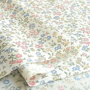 Può includere: Primo piano di tessuto floreale con sfondo crema. Il tessuto è ricoperto da piccoli e delicati fiori nei toni del rosa, blu, giallo e verde, con foglie verdi. Il tessuto è piegato, mostrando il motivo su entrambi i lati.