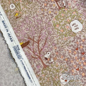 Può includere: Tessuto con un design vintage di Peanuts, con Snoopy, Woodstock ed elementi floreali. Il tessuto ha uno sfondo beige con illustrazioni rosa, verdi e marroni. Il testo "Vintage PEANUTS" e "MADE IN JAPAN" è visibile.