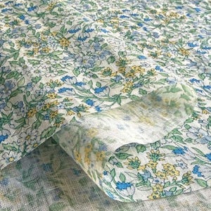 May include: Close-up of a floral fabric with a delicate pattern. The fabric features small blue, yellow, and white flowers with green leaves on a light background. The fabric is folded, showing the texture and layers of the material.