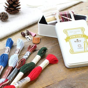 May include: A craft kit with embroidery floss in various colors, including blue, green, and red. The kit includes a white tin with the text "Enjoy weaving with POKEORI" and a DMC logo. Also included are small scissors and a leather thimble.
