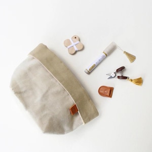 Puede incluir: Una bolsa de lona beige con una parte superior doblada, una etiqueta de madera en forma de mariposa, un marcador blanco, unas tijeras pequeñas y una etiqueta de cuero. Los artículos están dispuestos sobre una superficie blanca.