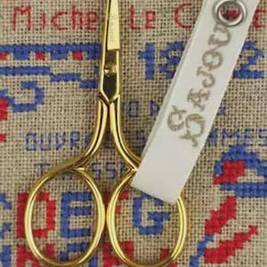 May include: Gold-colored embroidery scissors with a white tag that reads "Sajou". The scissors are positioned over a cross-stitch background with red, blue, and beige threads, creating a vintage aesthetic.