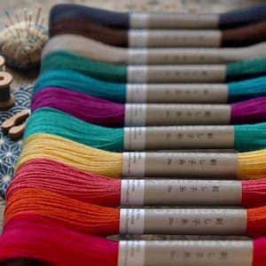 May include: A collection of embroidery thread skeins in various colors including dark blue, brown, cream, teal, purple, yellow, orange, and red. Each skein is wrapped in a paper band labeled with the text Sashiko and Japanese characters.