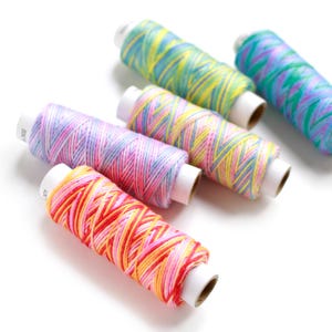 Puede incluir: Una colección de coloridas bobinas de hilo. Las bobinas están envueltas con hilos en varios tonos de rosa, amarillo, naranja, azul y verde. Las bobinas están dispuestas sobre un fondo blanco, creando una exhibición vibrante y artística.