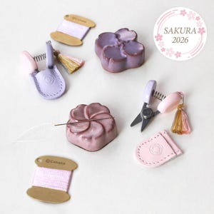 May include: A selection of pastel-coloured sewing tools and accessories. Includes small scissors, needle holders, thread spools, and flower-shaped items. The image also features the text "Sakura 2026" in a circular design.