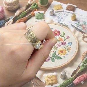 Puede incluir: Un dedal de latón con diseño floral, utilizado para bordar. El dedal está en un dedo, guiando hilo blanco hacia un bastidor de bordado con un diseño floral. Bobinas de hilo y suministros de bordado en el fondo.