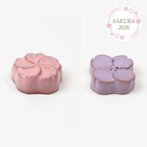 May include: Two flower-shaped confections, one pink and one lavender, sit on a white background. The pink flower has five petals, while the lavender one has four. A circular logo with the text "SAKURA 2026" is in the upper right corner.