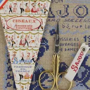 May include: A vintage sewing kit featuring gold scissors, a white Sajou tag, and a decorative box with the text "CISEAUX BRODERIE COUTURE" and illustrations of people. The kit is set against a beige and blue embroidered background.
