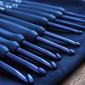 May include: A set of blue crochet hooks arranged on a dark blue fabric. The hooks have a metallic sheen and ergonomic handles. The hooks are angled towards the viewer, showcasing their curved tips. The image is well-lit, highlighting the tools' details.