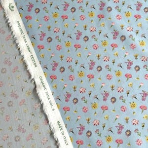 May include: Light blue organic cotton fabric with a floral pattern. The fabric features small, colorful flowers in shades of pink, yellow, and purple. The text "ORGANIC TANA LAWN COTTON" and "MADE IN ITALY" is visible on the fabric.