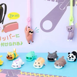 Könnte beinhalten: Illustration mit einem schwarzen Rucksack, einem rosa Federmäppchen und verschiedenen tierförmigen Reißverschlussanhängern. Die Reißverschlussanhänger zeigen ein Faultier, eine Katze und weitere Tiere in leuchtenden Farben.