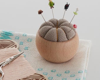 May include: A wooden pincushion with a brown, segmented top, holding decorative pins with miniature figures. The pincushion sits in a light-coloured wooden bowl. A pair of small scissors and spools of thread are in the foreground.