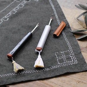 May include: Two seam rippers, one black and one silver, with wooden accents and tassels, are displayed on a dark gray embroidered fabric. A small brown leather sheath is also present. The tools are designed for sewing and crafting.