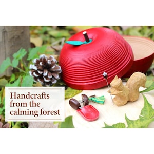 Puede incluir: Una caja de madera roja con forma de manzana con una hoja verde y un tallo negro, junto a una figura de ardilla de madera y una piña. El texto "Handcrafts from the calming forest" se muestra sobre un fondo blanco.