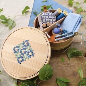 Pode incluir: Um bastidor de bordado de madeira com um desenho de ponto cruz azul e turquesa está ao lado de um kit de costura de madeira aberto. O kit contém carretéis de linha azul, uma almofada de alfinetes, tecido e outros utensílios de costura.