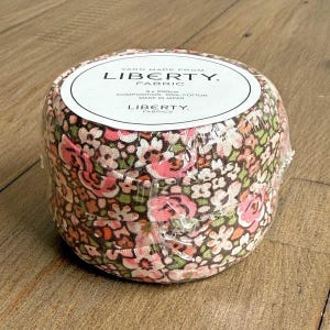 May include: A roll of Liberty Fabric, wrapped in clear plastic. The fabric features a floral pattern in pink, white, and green. The label reads "YARN MADE FROM LIBERTY. FABRIC" and "3 x 200cm COMPOSITION: 100% COTTON MADE IN JAPAN LIBERTY. FABRICS."