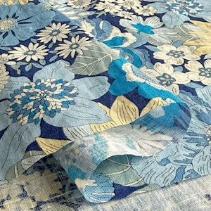Op de afbeelding: Close-up van een stof met een bloemenpatroon in tinten blauw, wit en geel. Het ontwerp toont grote, gestileerde bloemen en bladeren tegen een donkerblauwe achtergrond. De stof is gevouwen en onthult het patroon aan beide zijden.