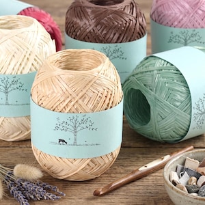 May include: Several balls of raffia yarn in various colors including beige, brown, pink, and green, each wrapped in a light blue paper band featuring a black line drawing of a tree and a small animal. A wooden crochet hook and dried lavender.