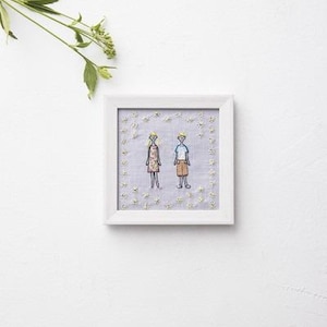 以下が含まれることがあります： 2人の様式化された人物が描かれた額入りの刺繍。一人は模様入りのドレス、もう一人は青いシャツとショートパンツを着ています。背景は、白いフレームの中に、刺繍された黄色の花が咲く水色の生地です。
